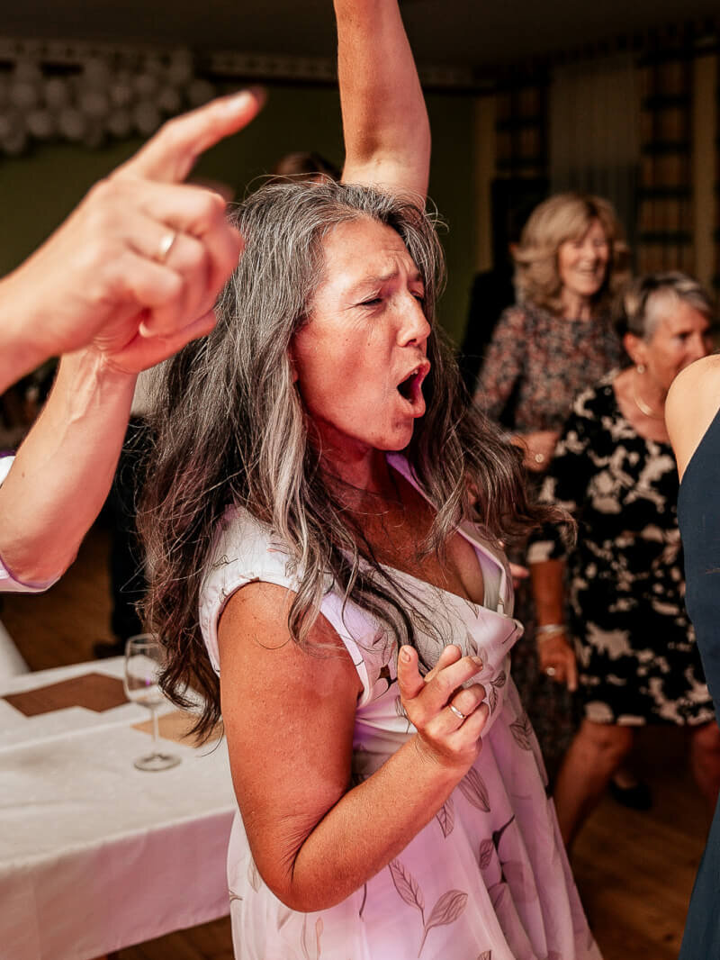 Eine Frau mit grauem Haar tanzt begeistert auf einer Party, gestikuliert und singt mit, umgeben von anderen fröhlichen Gästen in einer festlichen Innenatmosphäre.